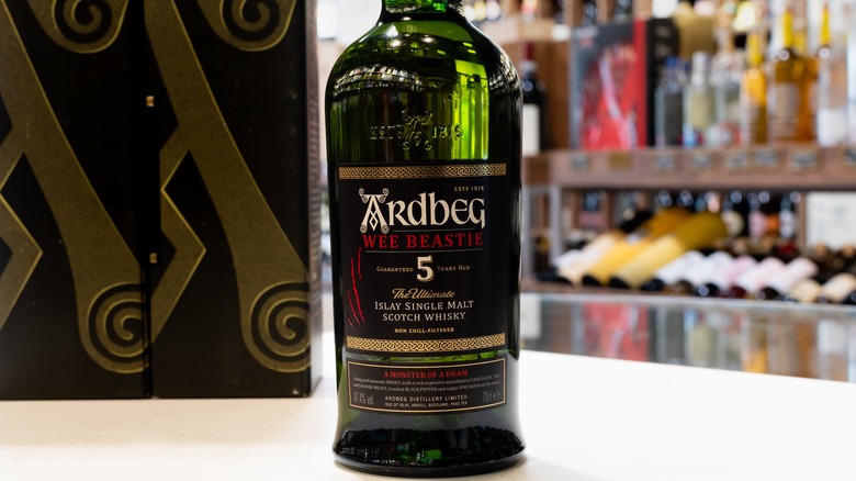 A bottle of Ardbeg Wee Beastie on a table with boxes with shelves of other bottles in the background
