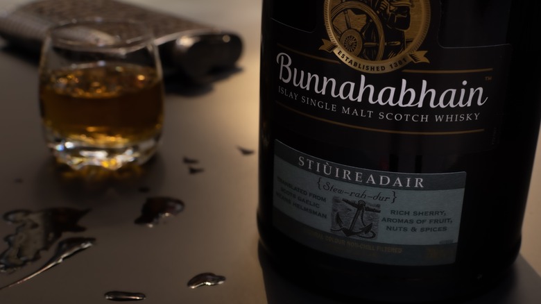 A close up of the bottom half of a bottle of Bunnahabhain Stiuireadir with a full glass and a whisky flask in the background