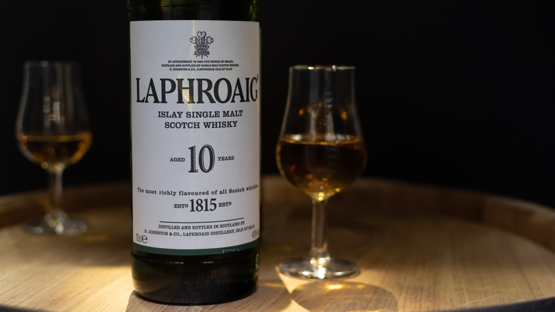 A bottle of Laphroaig 10-year on top of an upturned whisky barrel with full whisky glasses either side