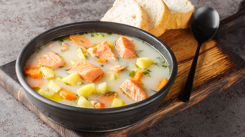 Bowl of fish chowder with chunks of salmon and diced potatoes sitting on top of cutting board with spoon and slices of bread nearby.