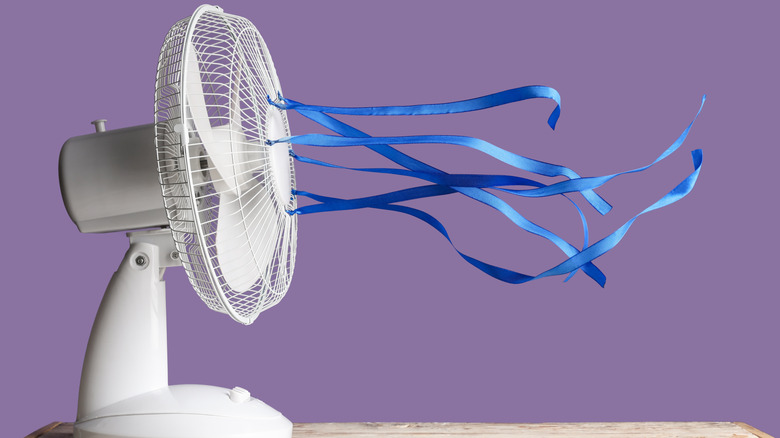 Electric fan with fluttering ribbons on table against violet background