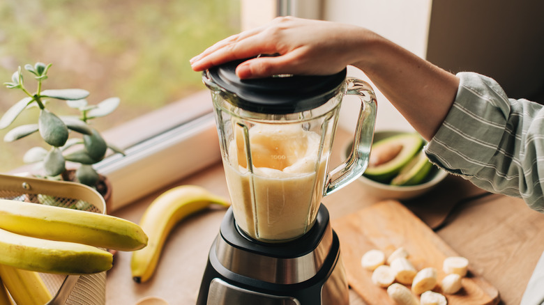 A hand on the lid of a blender filled with pureed banana on a counter by the window, with bananas in a basket and chopped bananas on a cutting board