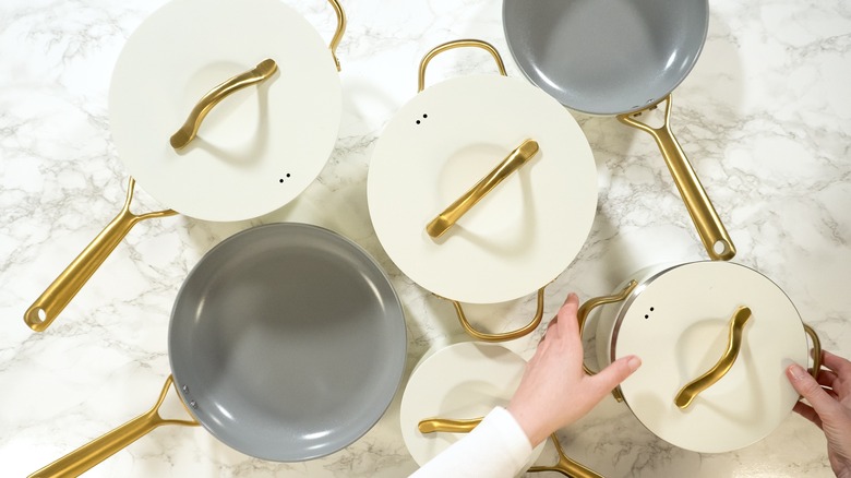 A set of white, gray, and gold pots and pans on a marbled countertop with female hands holding a lid
