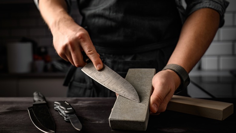 Chef sharpening knife on whetstone with other knives on table, in a dark kitchen
