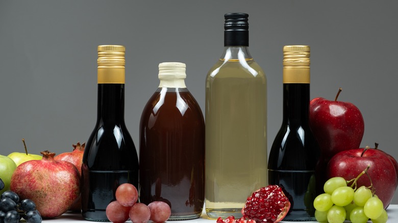 Different types of vinegars in clear bottles lined up with various fruits against a gray background