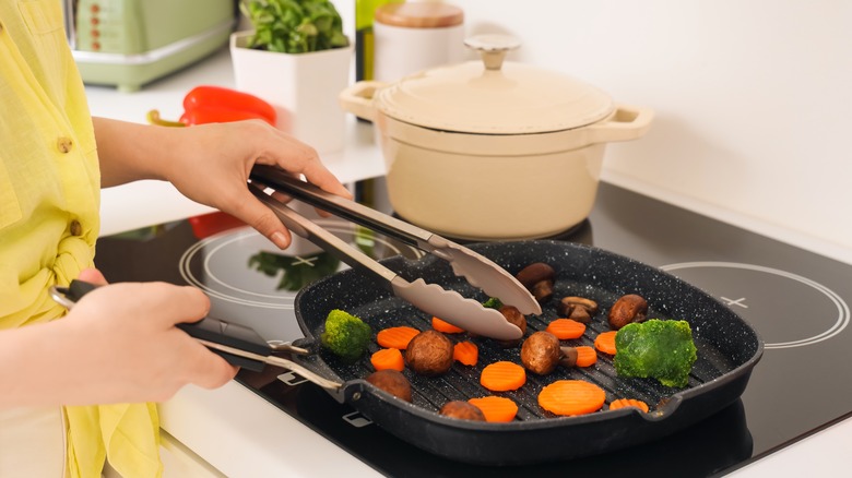 A woman using tongs to flip vegetables in a pan on the stove with other kitchen tools in the background
