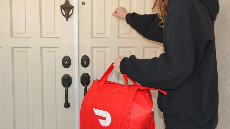 A DoorDash delivery person knocking on someone's door holding a red branded bag