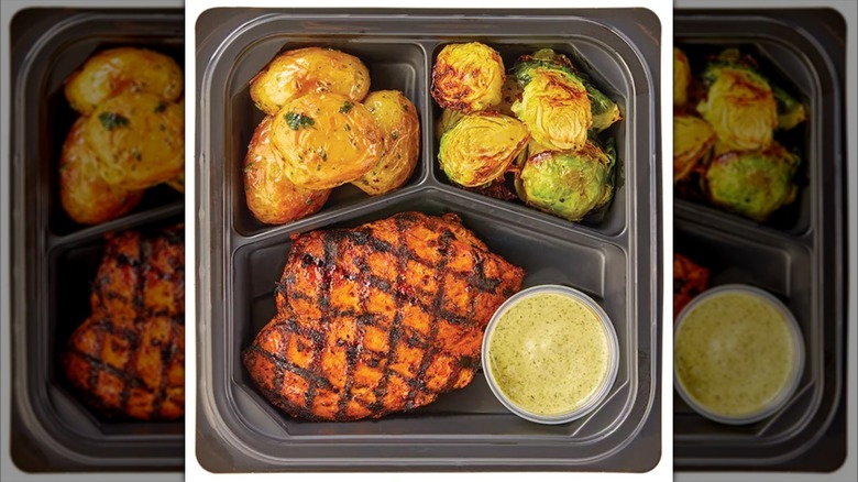 Top view of a meal tray with potatoes, grilled chicken, brussels sprouts, and sauce.