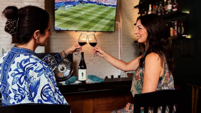 Two women clink wine glasses at a posh bar with sports on the TV