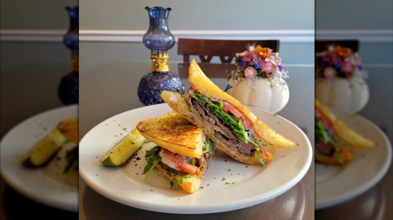 A prime rib panini on a white plate next to flowers and an old-fashioned oil lamp