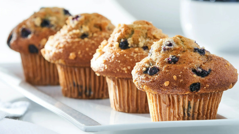 Lineup of four blueberry muffins on white plate with oranges in background