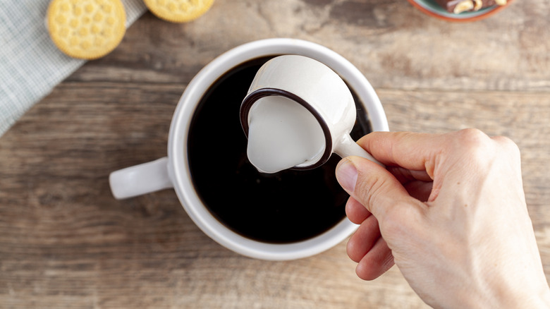 Top view of a person adding creamer to cup of black coffee