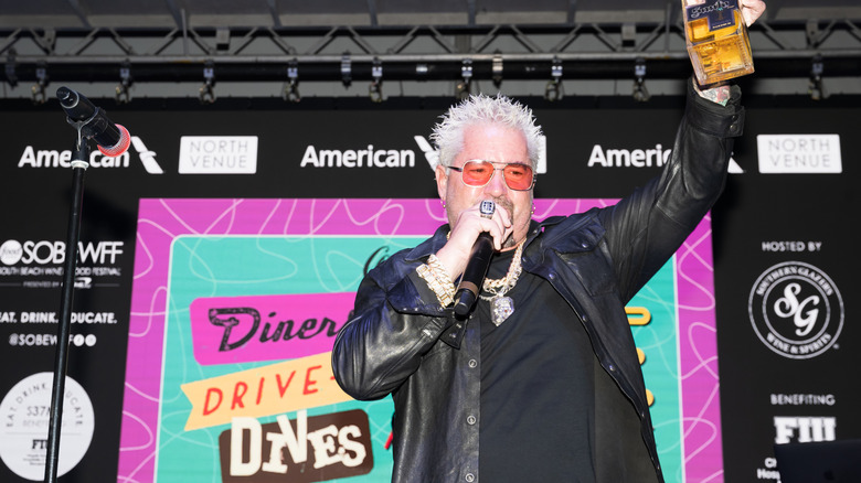 Guy Fieri holding microphone standing in front of Diners, Drive-Ins & Dives sign