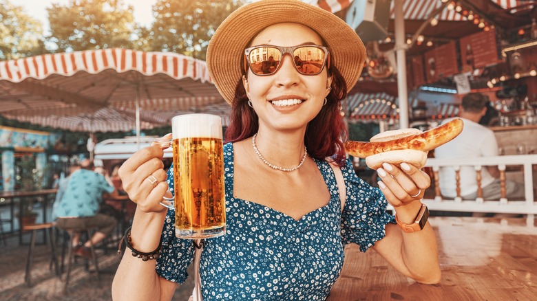 Smiling person wearing hat and sunglasses holding a beer and bratwurst