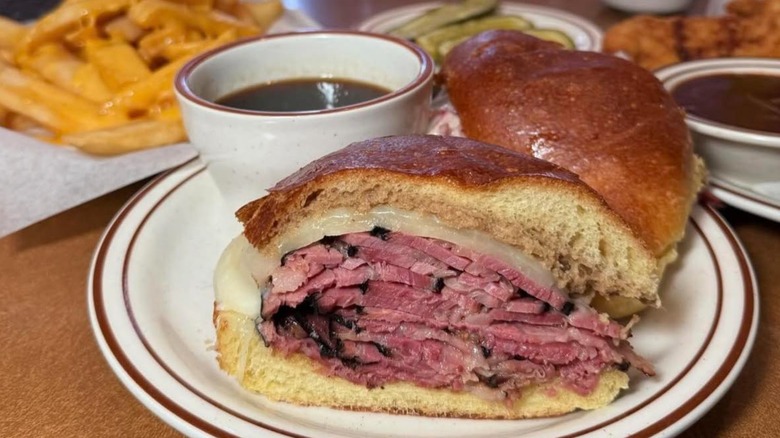 Sliced pastrami sandwich with cup of au jus and other dishes in background