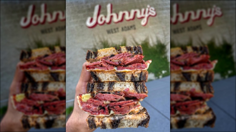 Hands holding two stacked halves of pastrami on marbled rye outside in front of Johnny's sign