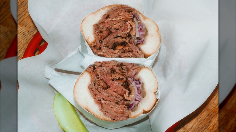 Two halves of pastrami sub wrapped in paper with pickle