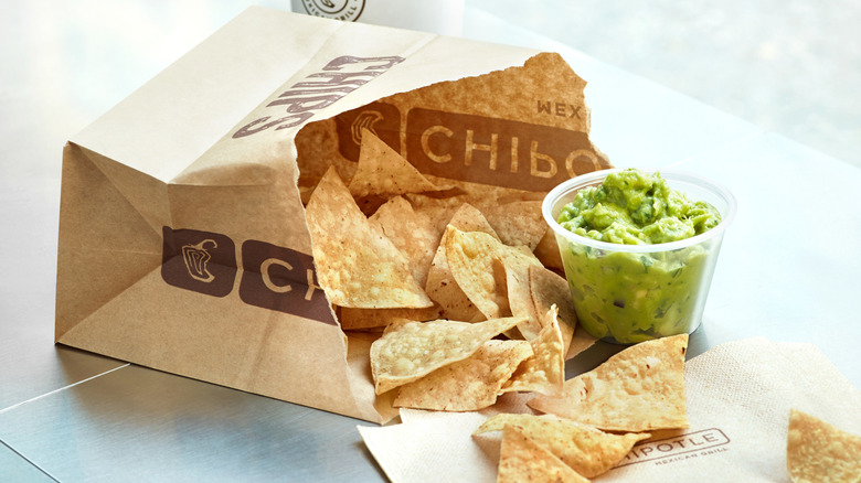A paper Chipotle bag with tortilla chips spilling out next to a small plastic container of guacamole