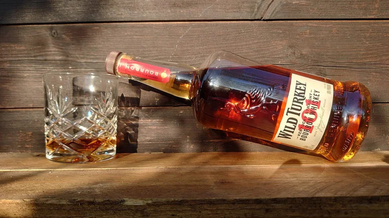 A bottle of Wild Turkey 101 resting against a filled glass on a wooden background