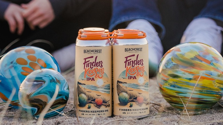 Two cans of Finders Keepers IPA in sand beside colorful glass floats