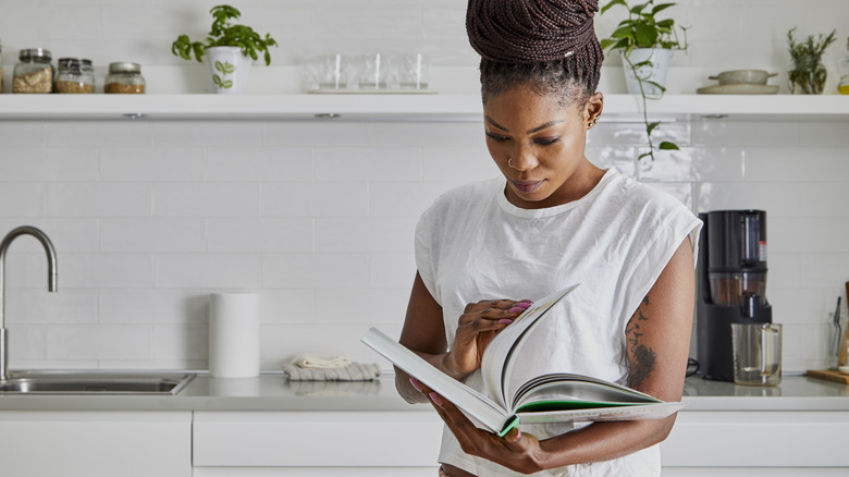 Woman standing in a kitchen reading a hardcover cookbook