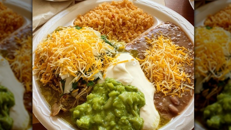White plate filled with rice, pork, and refried beans, topped with cheese, sour cream, and guacamole