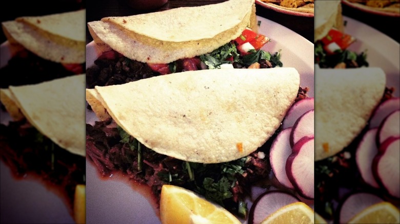 Two birria tacos on a plate with tomato, cilantro, radishes, and lemon wedges