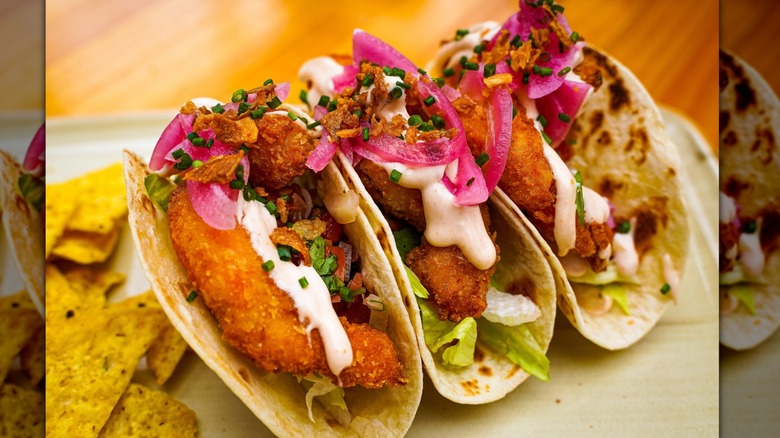 Three breaded and fried fish tacos served in fresh tortillas, topped with lettuce, onions, and a white sauce