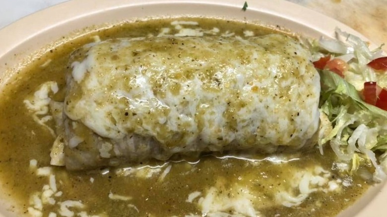 Large burrito on a white plate, smothered in a green sauce, and served with lettuce and tomato