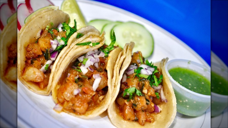 Three tacos topped with onions and cilantro, served with radishes, cucumbers, and a lime wedge
