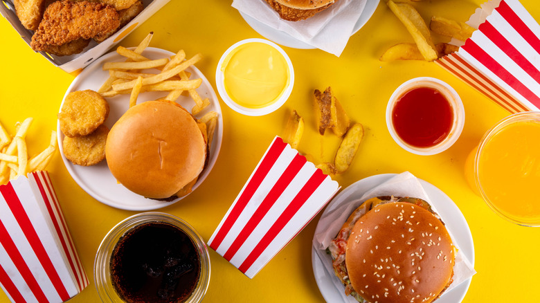 An assortment of fast foods on a yellow background including sodas, chicken tenders, burgers and fries