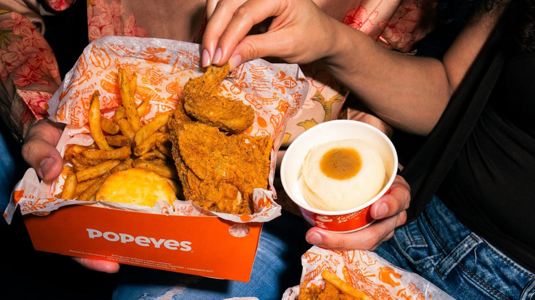 A Popeyes big box on two people's laps, with a biscuit, potatoes and gravy, fries and two pieces of chicken