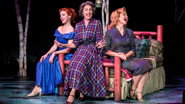 Three women in vintage dresses sitting on a bed on stage singing