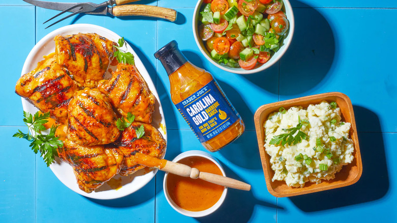 Bottle and bowl of Trader Joe's Carolina Gold Barbecue Sauce next to plate of grilled chicken, square bowl of potato salad, and bowl of tomatoes and cucumbers