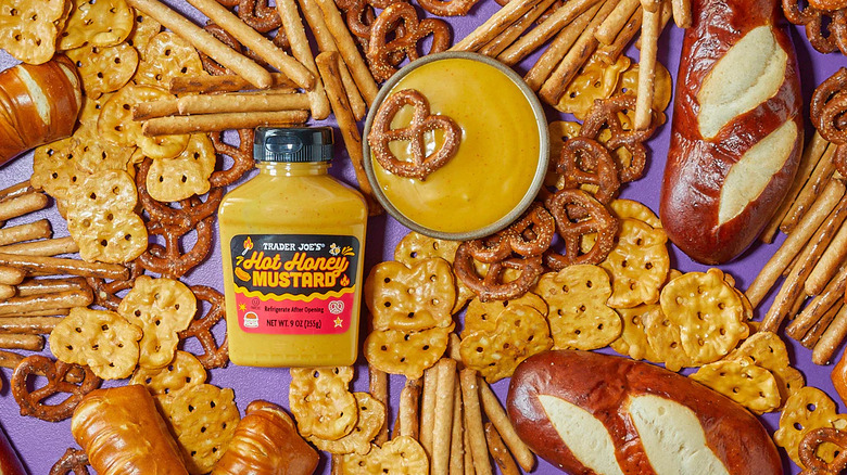 Bottle of Trader Joe's Hot Honey Mustard on purple table next to crackers, pretzels, and pretzel bread loaves