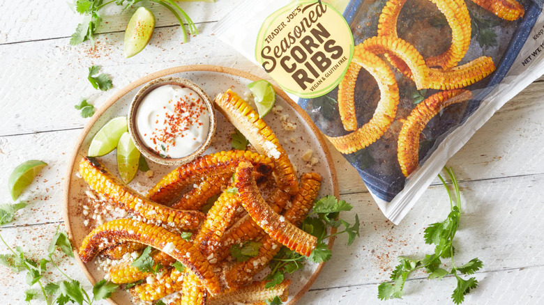 Bag of Trader Joe's Seasoned Corn Ribs next to plate of cooked corn ribs with dipping sauce, lime wedges, chili powder, and cilantro