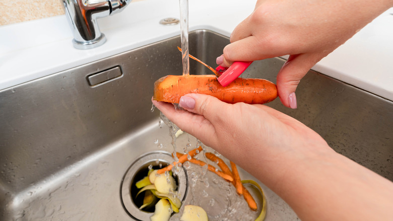 Hands peeling a fresh carrot under running water with waste collected in a garbage disposal