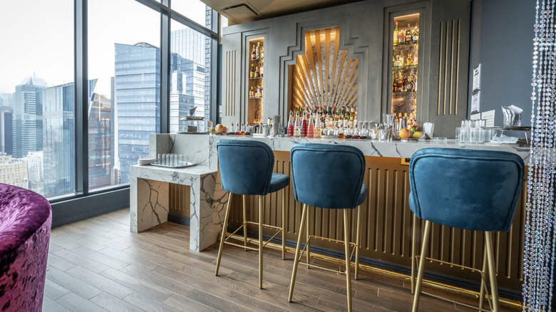A gold and white bar with blue bar chairs and a city view in the background.