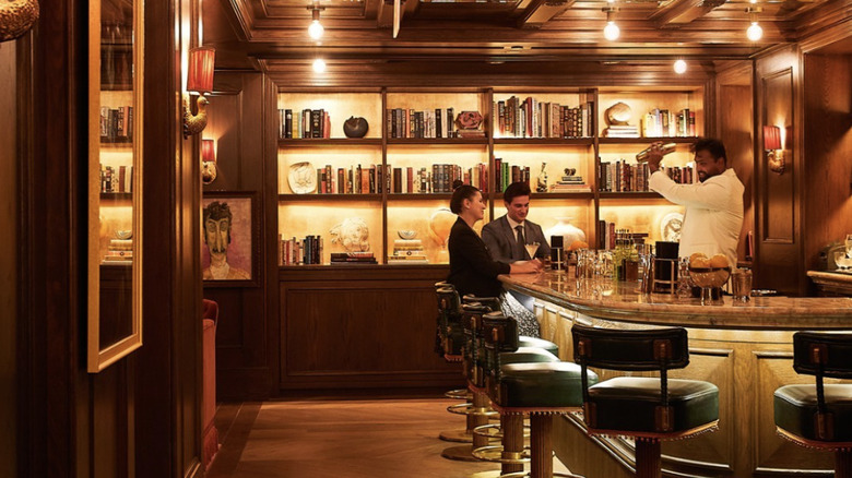 Two people sitting at a dimly lit bar while a bartender prepares a cocktail.