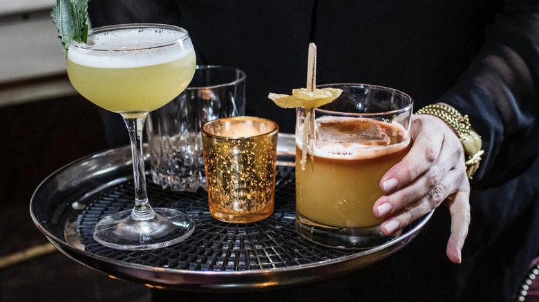 a person holding a silver tray with cocktails and a candle on top.