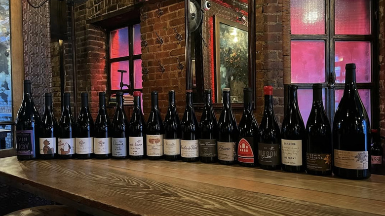 a line up of wine bottles on top of a wooden bar against a brick wall.