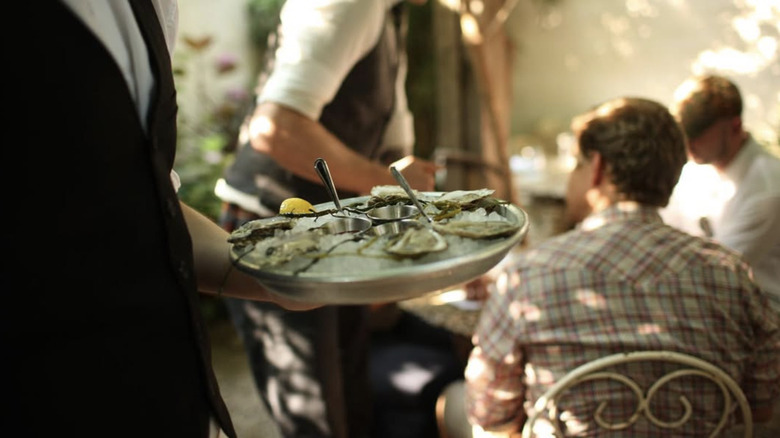 A server holding a dish of raw oysters with blurred diners in the background