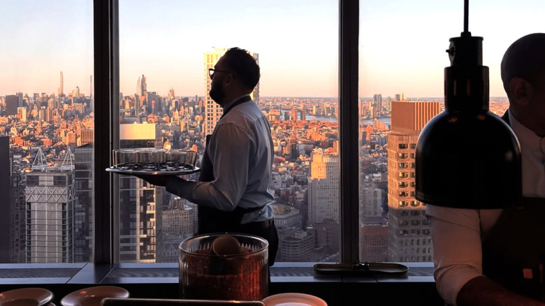 Server carrying a tray in restaurant with windows looking out at NYC in the backdrop