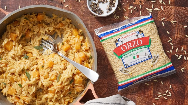Package of orzo on table beside pot of cooked pasta with vegetables and herbs