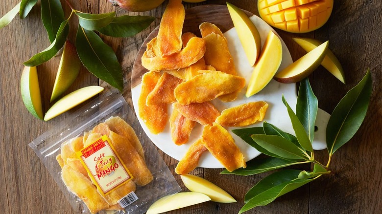 Package of dried mango on table alongside plate of dried and fresh mango and mango tree leaves
