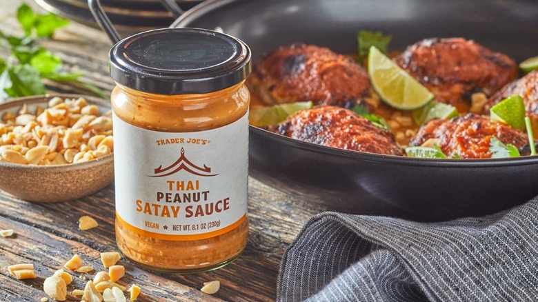 Glass jar of Thai peanut satay sauce in front of cooked meat entree and bowl of peanuts