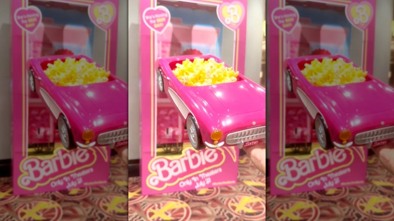 A person holds a pink "Barbie" popcorn bucket car in front of a large "Barbie" box