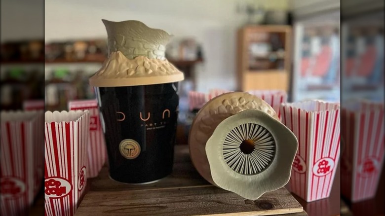 Two "Dune: Part Two" popcorn buckets sit, one on its side, on a wooden table surrounded by red and white popcorn boxes