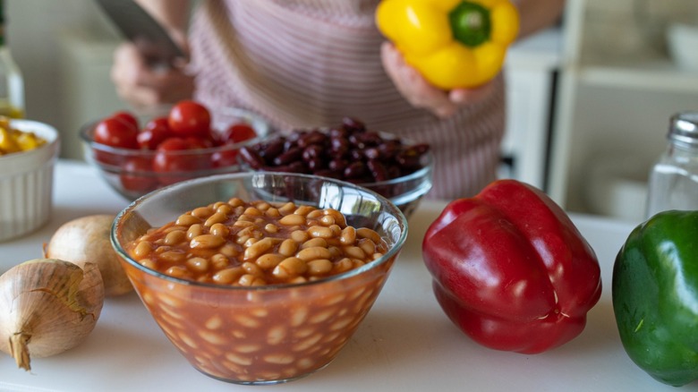 Baked beans in a bowl on a countertop next to peppers, kidney beans, onions, tomatoes