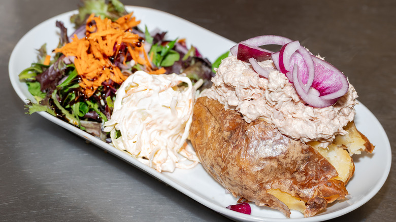 A baked potato topped with tuna salad and red onion next to some coleslaw and a mixed green salad with carrot